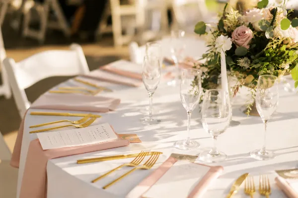 A close-up of a wedding reception table setting featuring white linens, gold cutlery, wine glasses, and a floral arrangement.