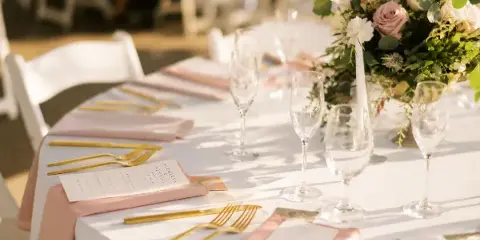 A close-up of a wedding reception table setting featuring white linens, gold cutlery, wine glasses, and a floral arrangement.