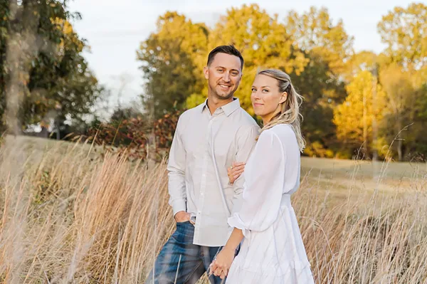 A smiling couple posing together in a field of tall grass with autumn trees in the background.