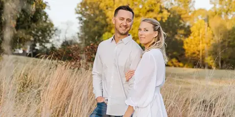 A smiling couple posing together in a field of tall grass with autumn trees in the background.