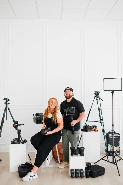 A man and a woman holding professional cameras and lenses in a studio with photography equipment.