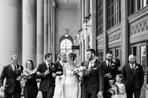 A black and white photograph of a bride and groom walking with their wedding party in a city street.