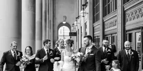 A black and white photograph of a bride and groom walking with their wedding party in a city street.
