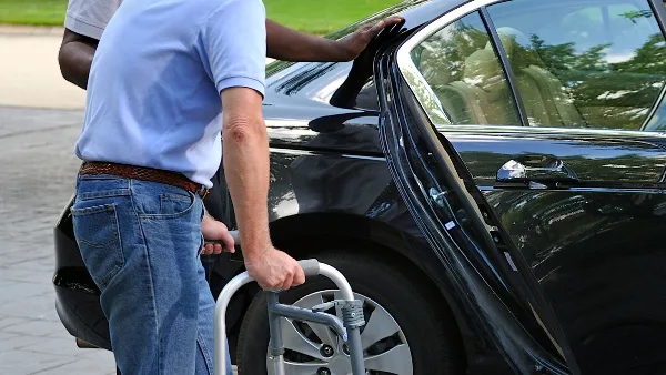 A driver assisting a passenger with a walker into a black sedan