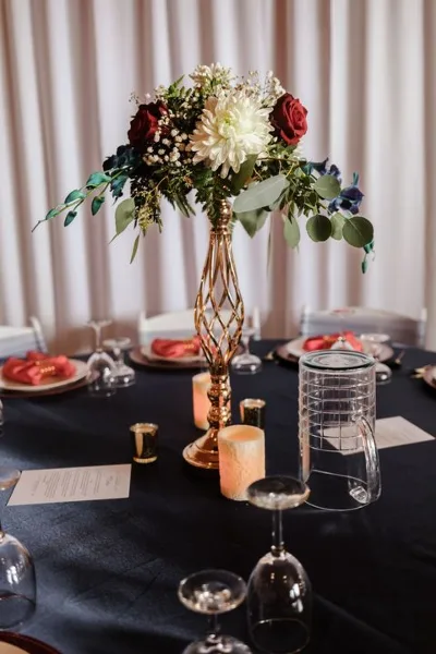 A decorated event table featuring a tall floral arrangement in a gold vase, candles, and glassware on a dark tablecloth.