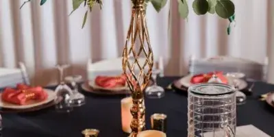 A decorated event table featuring a tall floral arrangement in a gold vase, candles, and glassware on a dark tablecloth.