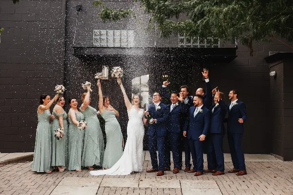 A wedding party, including the bride and groom, celebrating with champagne and confetti in front of a dark brick building.