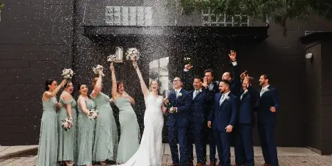 A wedding party, including the bride and groom, celebrating with champagne and confetti in front of a dark brick building.