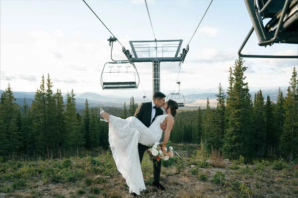 A bride and groom kissing in a mountainous landscape with a ski lift in the background.