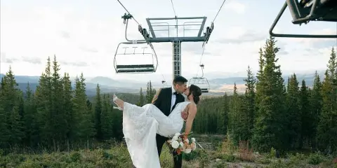 A bride and groom kissing in a mountainous landscape with a ski lift in the background.