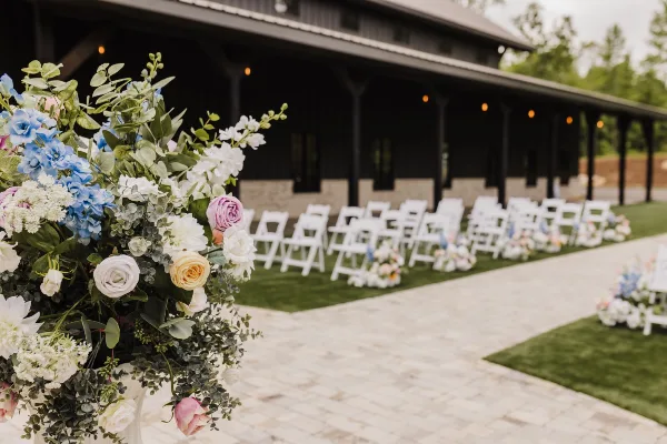 Outdoor wedding ceremony aisle with white chairs and floral arrangements set against a dark timber and stone venue building.