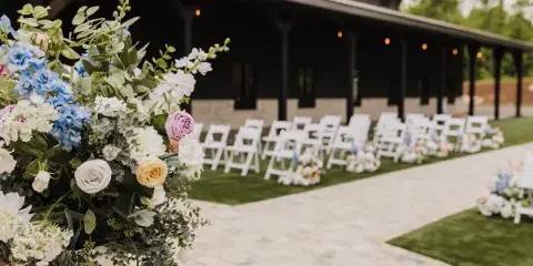 Outdoor wedding ceremony aisle with white chairs and floral arrangements set against a dark timber and stone venue building.