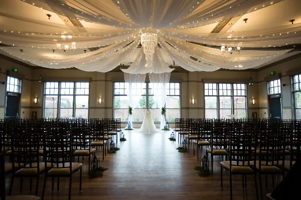 A wedding venue interior featuring a white wedding dress on a stand, rows of chairs, and a draped ceiling with a chandelier.
