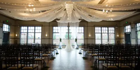 A wedding venue interior featuring a white wedding dress on a stand, rows of chairs, and a draped ceiling with a chandelier.