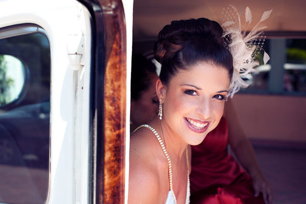 A smiling bride with an updo hairstyle and a white floral hair accessory, wearing a pearl necklace, looking out from a vehicle.