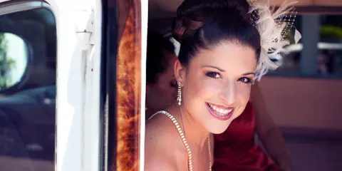 A smiling bride with an updo hairstyle and a white floral hair accessory, wearing a pearl necklace, looking out from a vehicle.