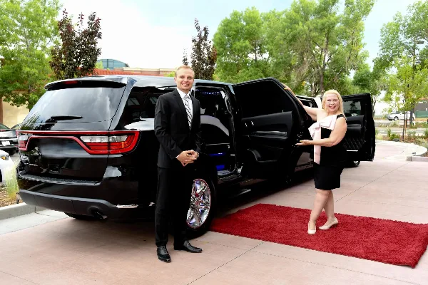 A professional chauffeur in a suit stands next to a black luxury SUV with the door open, while a woman steps out onto a red carpet.