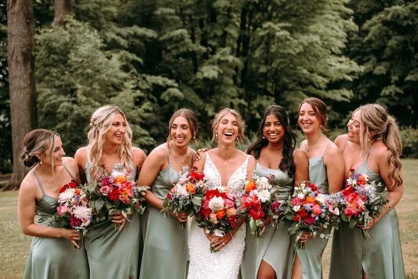 A bride and six bridesmaids in sage green dresses smiling and holding colorful floral bouquets outdoors.