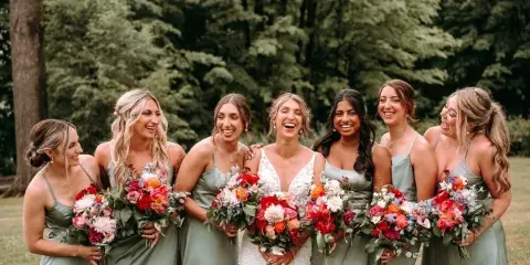 A bride and six bridesmaids in sage green dresses smiling and holding colorful floral bouquets outdoors.