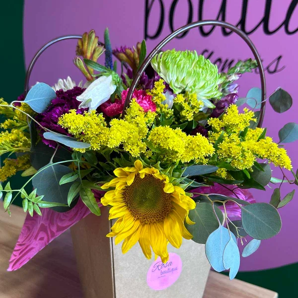 A mixed bouquet of sunflowers, yellow flowers, and purple blooms in a gift bag with a Rosa boutique sticker.