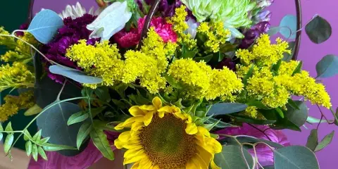 A mixed bouquet of sunflowers, yellow flowers, and purple blooms in a gift bag with a Rosa boutique sticker.