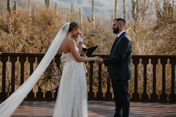A bride and groom holding hands during a wedding ceremony on a wooden deck with desert cacti in the background.