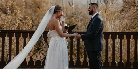 A bride and groom holding hands during a wedding ceremony on a wooden deck with desert cacti in the background.