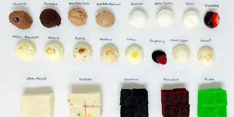 A cake tasting board with handwritten labels showing rows of filling and icing samples plus squares of white, vanilla, chocolate, red velvet, and pistachio cake.
