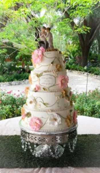 A four-tier white wedding cake decorated with pink and white sugar flowers and a couple topper, displayed on an ornate silver stand outdoors.