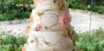 A four-tier white wedding cake decorated with pink and white sugar flowers and a couple topper, displayed on an ornate silver stand outdoors.