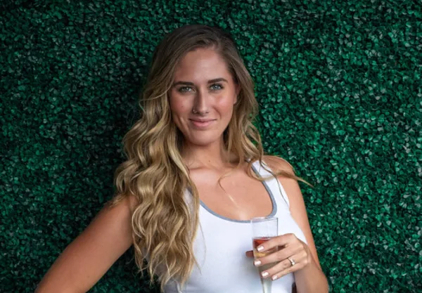 Portrait of a woman with styled wavy hair and makeup holding a glass against a green leafy background.