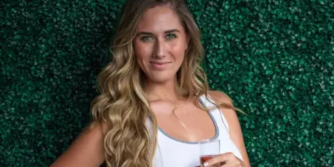Portrait of a woman with styled wavy hair and makeup holding a glass against a green leafy background.