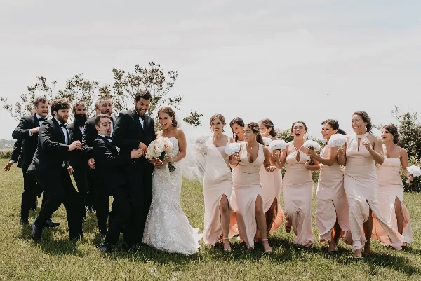 A bride and her wedding party, including groomsmen and bridesmaids, walking together in a grassy field.