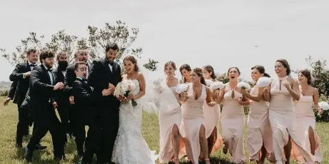 A bride and her wedding party, including groomsmen and bridesmaids, walking together in a grassy field.