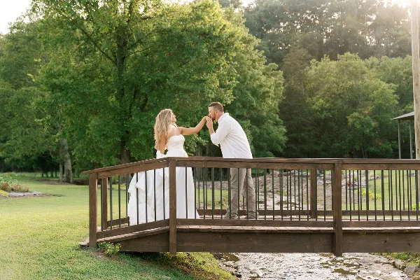 A bride and groom standing on a wooden footbridge over a creek in a wooded, rustic outdoor setting.