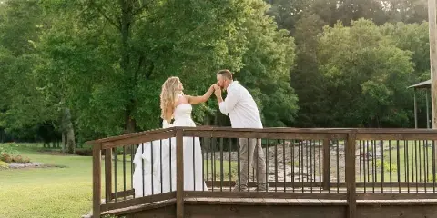 A bride and groom standing on a wooden footbridge over a creek in a wooded, rustic outdoor setting.