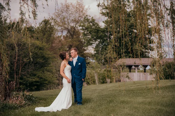 A bride in a white gown and a groom in a blue suit standing together in a grassy field with trees in the background.