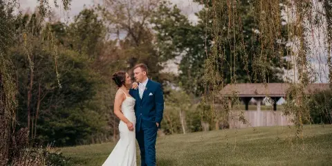 A bride in a white gown and a groom in a blue suit standing together in a grassy field with trees in the background.