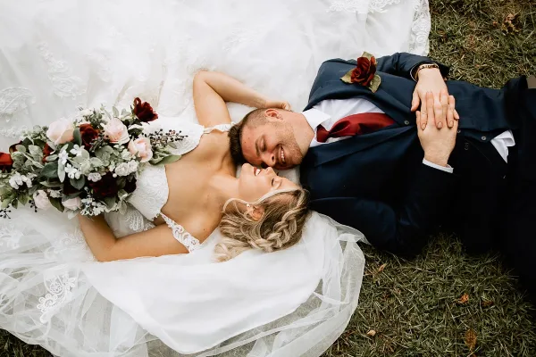 A bride and groom lying on the grass, smiling at each other, with the bride holding a bouquet.