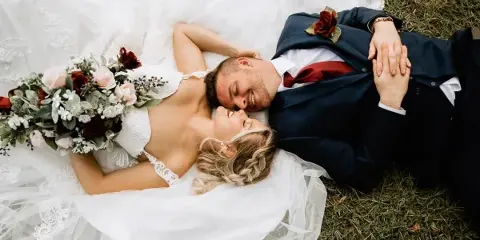 A bride and groom lying on the grass, smiling at each other, with the bride holding a bouquet.
