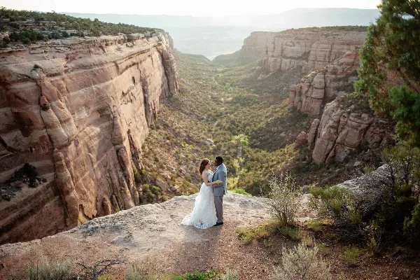 A bride and groom embracing on a rocky cliff edge overlooking a deep canyon with red rock formations.