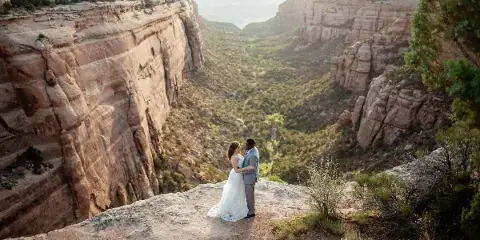 A bride and groom embracing on a rocky cliff edge overlooking a deep canyon with red rock formations.
