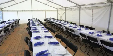 Interior of a large white event tent set up with long banquet tables, black chairs, and blue napkins.