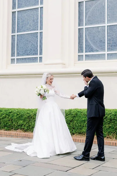 A bride in a white lace gown and a groom in a black suit holding hands and laughing in front of a building with stained glass windows.