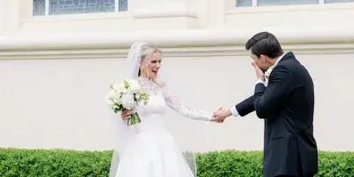 A bride in a white lace gown and a groom in a black suit holding hands and laughing in front of a building with stained glass windows.