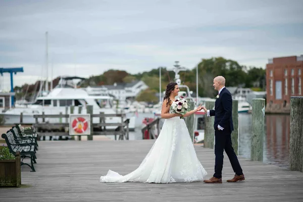 A bride and groom holding hands on a wooden dock with boats in the background.