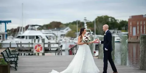 A bride and groom holding hands on a wooden dock with boats in the background.