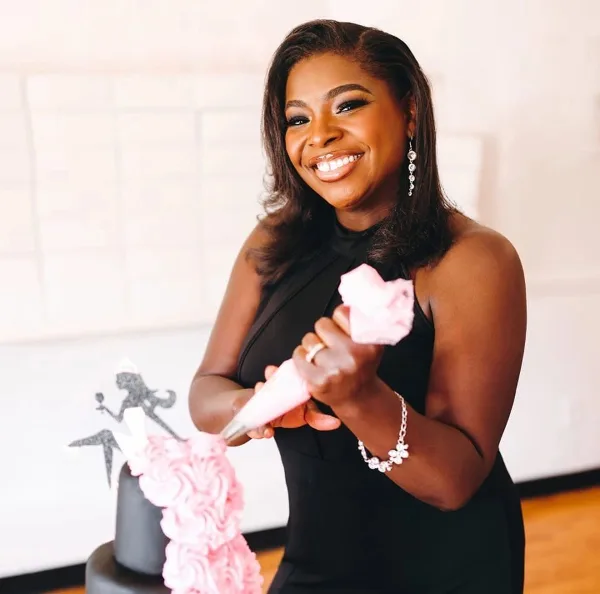 A smiling woman in a black dress piping pink frosting onto a cake.