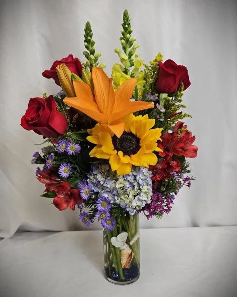 A vibrant floral arrangement with red roses, an orange lily, and a sunflower in a glass vase.