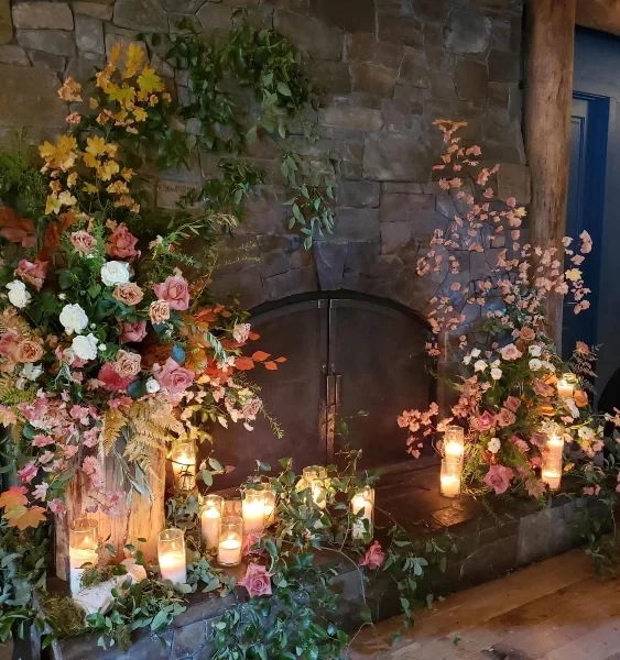 A stone fireplace decorated with lush floral arrangements, greenery, and lit candles.